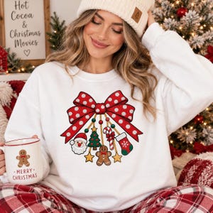 May include: A white long-sleeve sweatshirt featuring a large red polka-dot bow graphic with hanging Christmas ornaments including Santa, a tree, a candy cane, a stocking, and a gingerbread man. A matching white mug displays a gingerbread man.