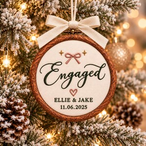 May include: A round wooden embroidery hoop ornament featuring cream fabric with the embroidered text Engaged, Ellie & Jake, and 11.06.2025. The design includes a pink stitched bow, a small pink heart, two gold stars, and a cream ribbon bow on top.