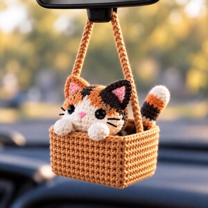 May include: A crocheted calico cat ornament sitting inside a small tan crocheted basket. The cat has white, orange, and dark brown fur, black eyes, and a pink nose. The basket features a long tan handle for hanging from a rearview mirror.