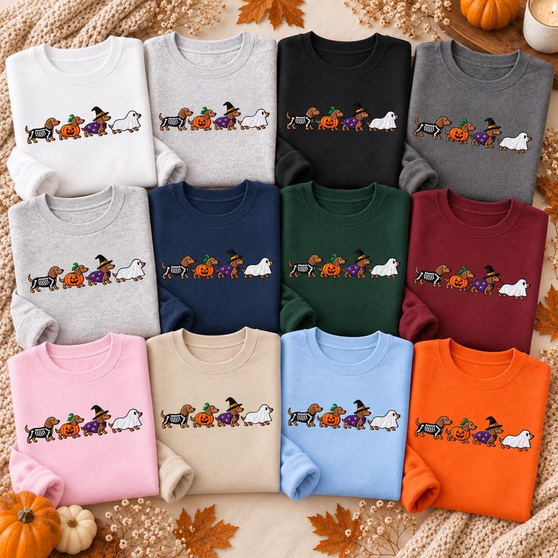 May include: A collection of twelve crewneck sweatshirts in various colors, each featuring a graphic of four dachshunds in Halloween costumes: one as a skeleton, one inside a pumpkin, one as a witch, and one dressed as a ghost.