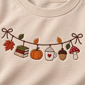 May include: A light beige sweatshirt featuring an embroidered garland design. The garland is suspended by two brown bows and displays a stack of books, an orange pumpkin, a white mug with a red heart, an acorn, and a red and white mushroom.