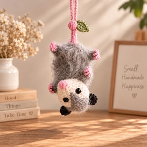 May include: A crocheted hanging ornament shaped like a fuzzy gray and white opossum. The creature has a pink nose, small pink paws, black ears, and black eyes. It hangs from a pink crocheted cord topped with a small green crocheted leaf.