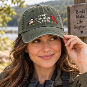 May include: An olive green baseball cap featuring an embroidered design of a black-capped chickadee and a red cardinal perched on branches. The text "easily distracted by birds" is embroidered in white lettering between the two bird illustrations.
