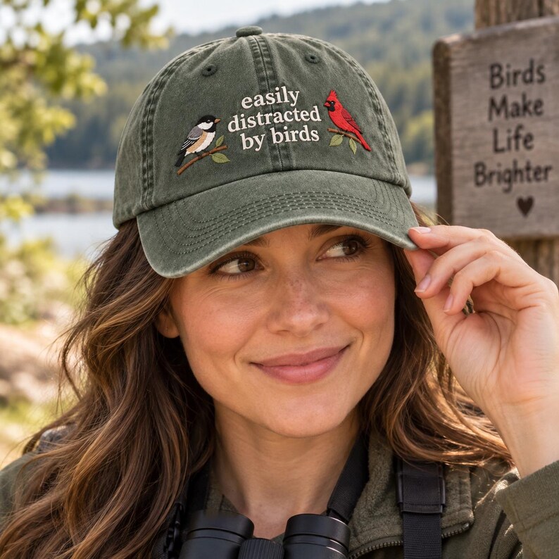 May include: An olive green baseball cap featuring an embroidered design of a black-capped chickadee and a red cardinal perched on branches. The text "easily distracted by birds" is embroidered in white lettering between the two bird illustrations.