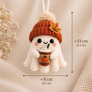 May include: A crocheted ghost ornament wearing a burnt orange beanie with a maple leaf accent, holding a coffee cup with a pumpkin design. The figure has black eyes and pink cheeks. It measures approximately 5.1 inches tall and 3.1 inches wide.