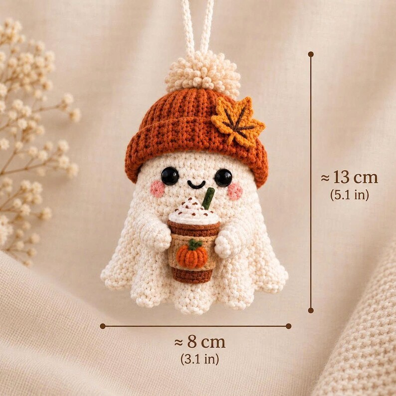 May include: A crocheted ghost ornament wearing a burnt orange beanie with a maple leaf accent, holding a coffee cup with a pumpkin design. The figure has black eyes and pink cheeks. It measures approximately 5.1 inches tall and 3.1 inches wide.