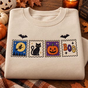 May include: A beige crewneck sweatshirt featuring four embroidered rectangular patches resembling postage stamps. The designs include a witch on a broomstick, a black cat, a jack-o'-lantern, and the word BOO, accented by two small black bats.