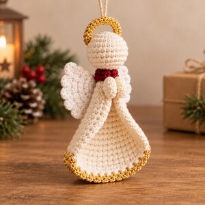 May include: A crocheted angel ornament in cream yarn with a gold halo and gold trim at the base of the dress. The figure features a small red collar, scalloped wings, and hands clasped together in front. A light-colored string is attached to the top.