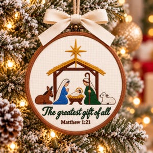 May include: A round wooden embroidery hoop ornament featuring a nativity scene with a donkey, Mary, baby Jesus in a manger, Joseph, and a lamb under a gold star. The text reads The greatest gift of all Matthew 1:21. A cream ribbon bow is at the top.