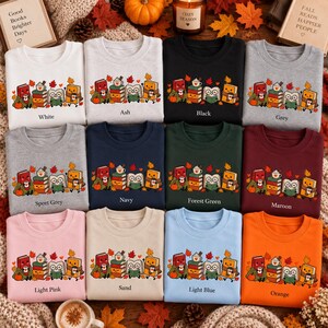 May include: A collection of twelve crewneck sweatshirts in various colors, each featuring a graphic of four anthropomorphic books with faces, autumn leaves, and a mug of hot cocoa. The text labels include White, Ash, Black, Grey, Sport Grey, Navy, Forest Green, Maroon, Light Pink, Sand, Light Blue, and Orange.