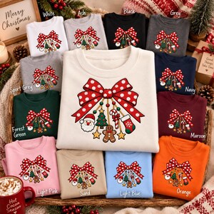 May include: A collection of crewneck sweatshirts in various colors, each featuring a central graphic of a large red and white polka dot bow with hanging Christmas ornaments including a Santa face, a tree, a candy cane, and a gingerbread man.