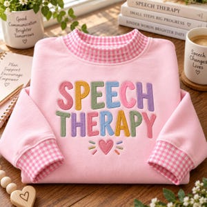 May include: A light pink sweatshirt featuring a pink and white gingham collar and cuffs. The front displays the embroidered text SPEECH THERAPY in multi-colored letters, with a small pink heart and radiating lines embroidered below the text.