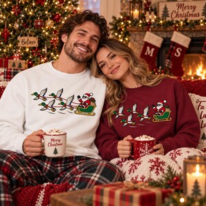 May include: A white crewneck sweatshirt and a maroon crewneck sweatshirt, both featuring a graphic of Santa Claus in a sleigh pulled by a team of flying mallard ducks. Each sweatshirt is paired with a ceramic mug containing a hot beverage.
