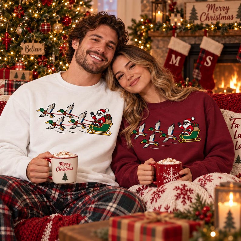 May include: A white crewneck sweatshirt and a maroon crewneck sweatshirt, both featuring a graphic of Santa Claus in a sleigh pulled by a team of flying mallard ducks. Each sweatshirt is paired with a ceramic mug containing a hot beverage.