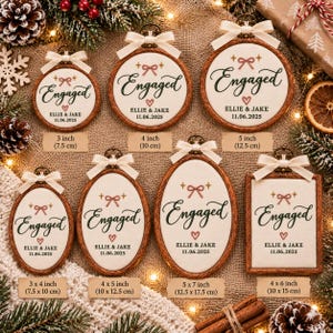 May include: A collection of wooden-framed embroidery hoops and a rectangular frame featuring white fabric with the embroidered text Engaged, the names Ellie & Jake, and the date 11.06.2025, accented with a pink bow, heart, and cream ribbon.