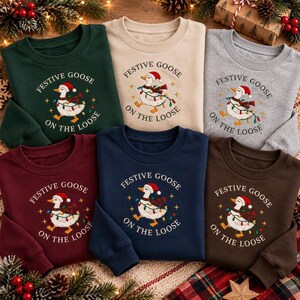 May include: A collection of six crewneck sweatshirts in dark green, beige, heather gray, maroon, navy blue, and dark brown. Each features a central graphic of a white goose wearing a Santa hat and string lights, with the text Festive Goose On The Loose.