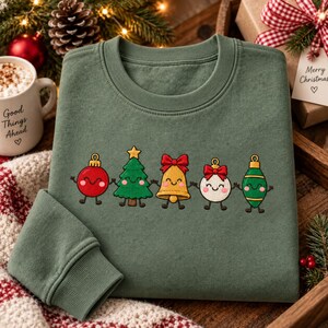 May include: An olive green crewneck sweatshirt featuring an embroidered graphic of five festive characters holding hands: a red ornament, a green Christmas tree, a gold bell, a white ornament with a red bow, and a green teardrop ornament.