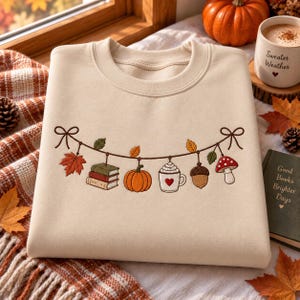 May include: A beige crewneck sweatshirt featuring an embroidered garland design across the chest. The garland includes a stack of books, a pumpkin, a mug with a heart and whipped cream, an acorn, and a mushroom, all accented by autumn leaves.