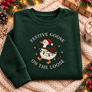 May include: A dark green crewneck sweatshirt featuring a graphic of a white goose wearing a red and white Santa hat and a red and green scarf. The goose is wrapped in colorful Christmas lights. The text reads FESTIVE GOOSE ON THE LOOSE.