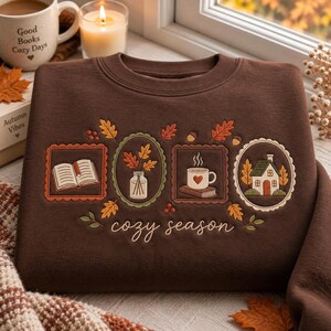 May include: A dark brown crewneck sweatshirt featuring embroidered designs of an open book, a vase with autumn leaves, a steaming mug with a heart, and a small house. The text "cozy season" is embroidered in white cursive below the designs.