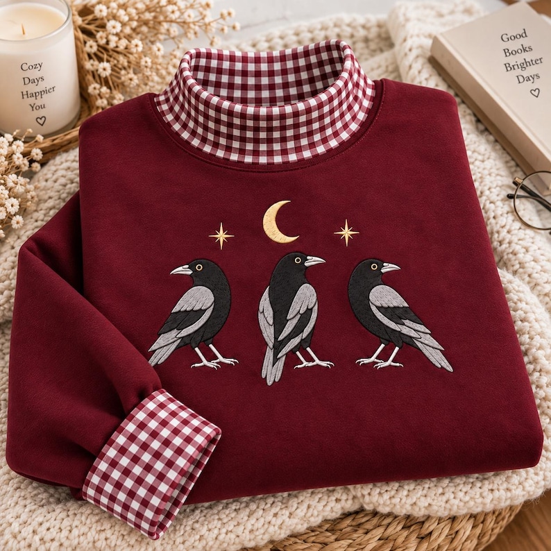May include: A deep maroon long-sleeve sweatshirt featuring a high mock neck collar and sleeve cuffs in a red and white gingham pattern. The chest displays an embroidered design of three black crows positioned beneath a gold crescent moon and stars.