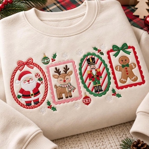 May include: A cream-colored crewneck sweatshirt featuring embroidered Christmas motifs. The design includes a Santa Claus in a red oval frame, a reindeer in a pink rectangular frame, a nutcracker in a green oval frame, and a gingerbread man in a red rectangular frame.