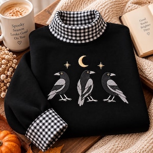 May include: A black long-sleeve sweatshirt featuring a mock neck collar and sleeve cuffs in a black and white gingham pattern. The chest displays an embroidered design of three black crows positioned beneath a gold crescent moon and two stars.