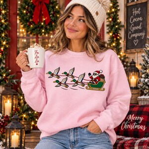 May include: A light pink crewneck sweatshirt featuring a graphic of Santa Claus riding in a green sleigh pulled by four mallard ducks. The sweatshirt is worn with a cream-colored knit beanie. A white mug with the red text Fa La La is held.