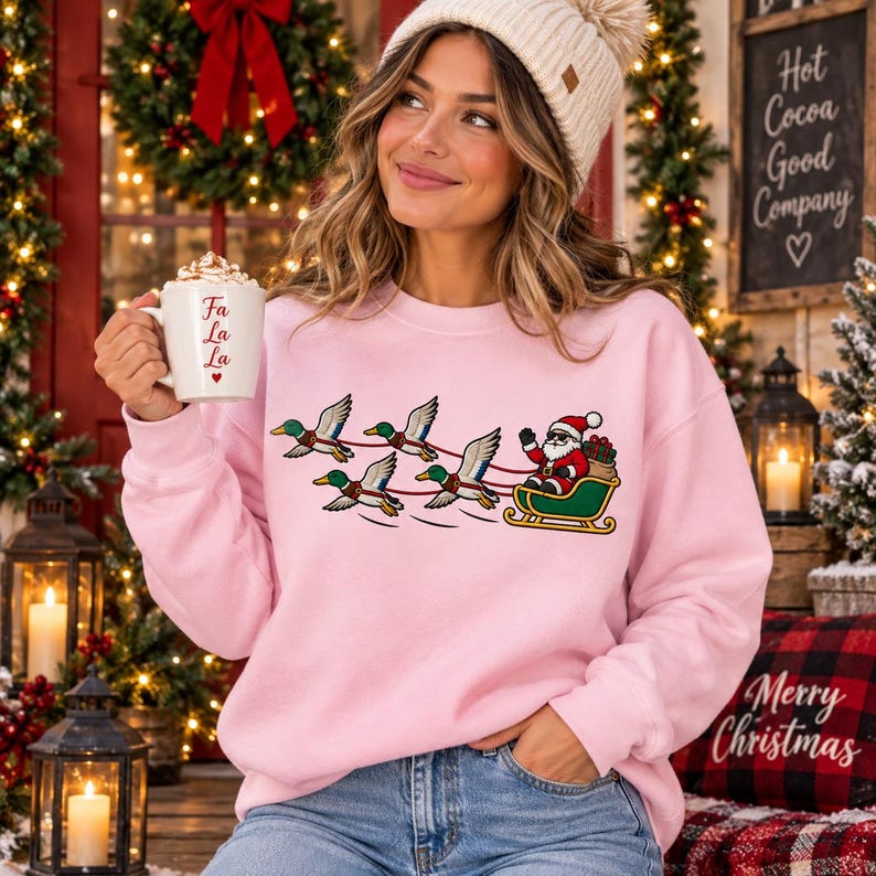 May include: A light pink crewneck sweatshirt featuring a graphic of Santa Claus riding in a green sleigh pulled by four mallard ducks. The sweatshirt is worn with a cream-colored knit beanie. A white mug with the red text Fa La La is held.