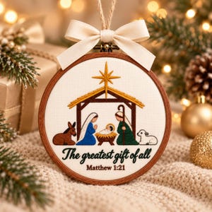 May include: A circular wooden embroidery hoop ornament featuring a nativity scene with a gold star, a donkey, Mary, Joseph, a baby in a manger, and a lamb. The text reads, "The greatest gift of all" and "Matthew 1:21". A cream ribbon bow is attached.