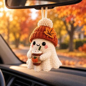 May include: A crocheted ghost ornament hanging from a rearview mirror. The white ghost wears an orange knit beanie with a brown maple leaf accent and holds a brown coffee cup featuring a small orange pumpkin graphic and a green straw.