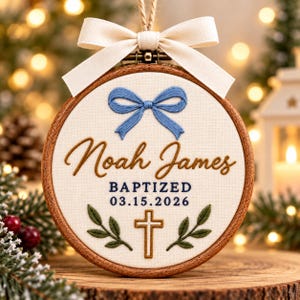May include: A circular wooden embroidery hoop containing off-white fabric with a blue embroidered bow, the name Noah James, the text BAPTIZED 03.15.2026, a cross, and green leaves. A cream-colored fabric ribbon is tied to the top of the hoop.