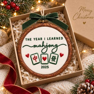 May include: A round wooden embroidery hoop ornament featuring white fabric with green embroidered text that reads THE YEAR I LEARNED mahjong 2025, three mahjong tiles, and two red hearts, topped with a dark green glittery fabric bow.
