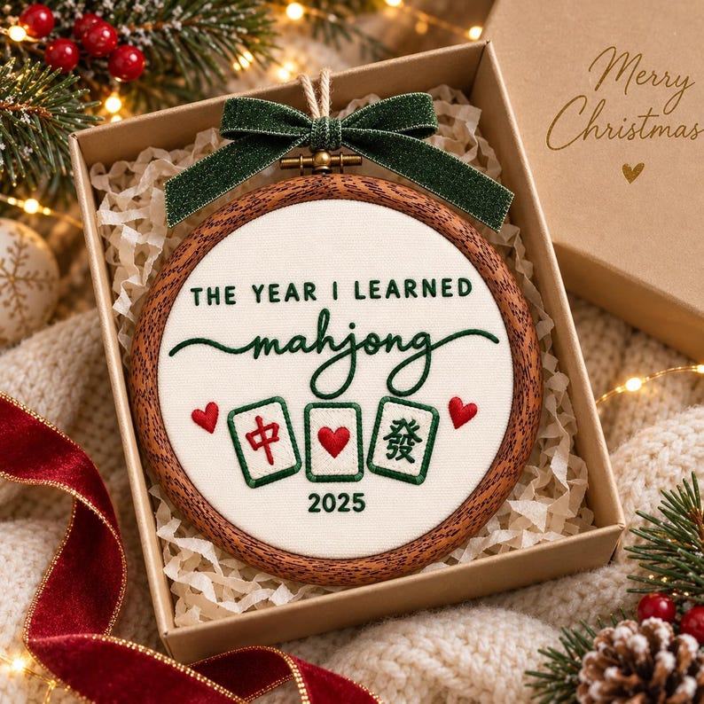 May include: A round wooden embroidery hoop ornament featuring white fabric with green embroidered text that reads THE YEAR I LEARNED mahjong 2025, three mahjong tiles, and two red hearts, topped with a dark green glittery fabric bow.