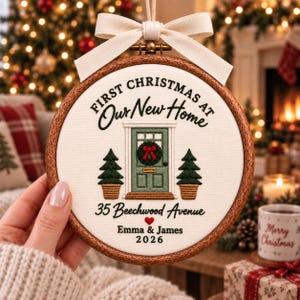 May include: A round wooden embroidery hoop ornament featuring white fabric with embroidered text First Christmas at Our New Home, 35 Beechwood Avenue, Emma & James, and 2026. It depicts a green door with a wreath and two potted pine trees.