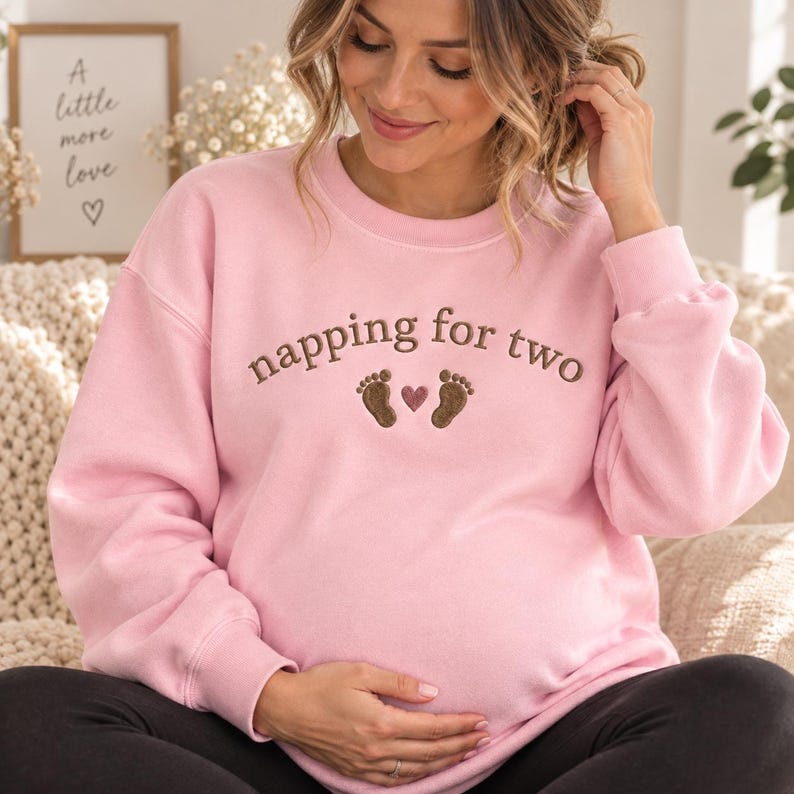 May include: A light pink crewneck sweatshirt featuring brown embroidered text that reads napping for two, accompanied by two small brown baby footprints and a small pink heart centered between the footprints on the front of the garment.