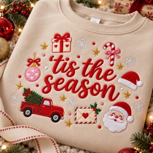May include: A beige crewneck sweatshirt featuring red embroidered text that reads 'tis the season'. The design includes a red truck carrying a green Christmas tree, a gift box, a candy cane, a Santa Claus face, an envelope, and snowflakes.