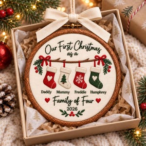 May include: A round wooden embroidery hoop ornament featuring cream fabric with embroidered text Our First Christmas as a Family of Four 2026 and four stockings labeled Daddy Mummy Freddie and Humphrey with red and green holiday accents.