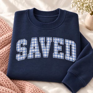 May include: A navy blue crewneck sweatshirt featuring the word SAVED in large, block-style letters. The letters are filled with a light blue and white gingham check pattern and are outlined with white stitching against the dark fabric.