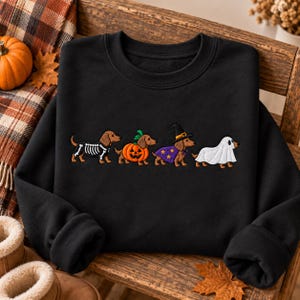 May include: A black crewneck sweatshirt featuring an embroidered design of four dachshunds in Halloween costumes: one in a skeleton suit, one inside a pumpkin, one wearing a witch hat and purple star-patterned cape, and one dressed as a ghost.