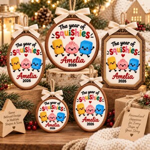 May include: Five embroidered ornaments in various shapes featuring the text The year of Squishies, the name Amelia, and the year 2026. Each piece displays a yellow rectangle, a pink dumpling, and a blue square character, topped with a cream bow.