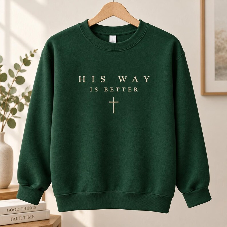 May include: A dark forest green crewneck sweatshirt featuring the text "HIS WAY IS BETTER" printed in a light cream color on the chest, positioned above a simple, thin, vertical cross symbol. The garment has long sleeves and ribbed cuffs.