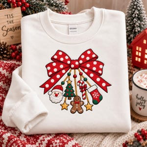 May include: A white crewneck sweatshirt featuring a large red bow with white polka dots. Hanging from the bow are gold chains with Christmas charms including a Santa Claus face, a green tree, a candy cane, a stocking, and yellow stars.