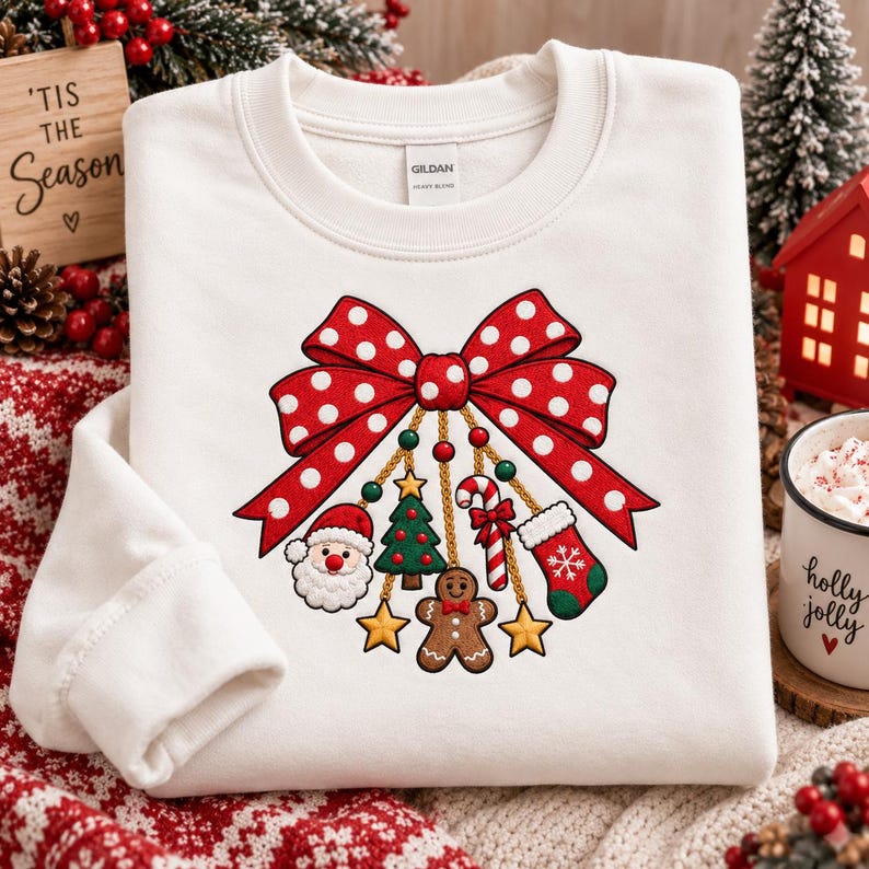 May include: A white crewneck sweatshirt featuring a large red bow with white polka dots. Hanging from the bow are gold chains with Christmas charms including a Santa Claus face, a green tree, a candy cane, a stocking, and yellow stars.