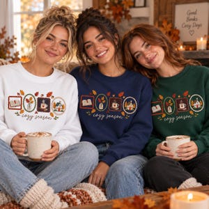 May include: Three crewneck sweatshirts in white, navy blue, and dark green, each featuring a graphic print of four framed autumn-themed icons including a book, leaves, a mug, and a house, with the text cozy season written in cursive script.