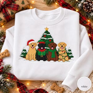 May include: A white crewneck sweatshirt featuring an embroidered graphic of four dogs sitting in front of a Christmas tree. The dogs include a yellow Labrador in a Santa hat, a black Labrador in an elf outfit, a chocolate Labrador in a green scarf, and a yellow Labrador wrapped in colorful string lights.