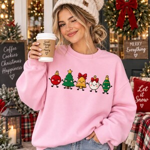 May include: A light pink crewneck sweatshirt featuring a graphic print of five cartoon Christmas ornaments holding hands, including a red bauble, a green tree, a yellow bell, a white bauble with a red bow, and a green teardrop ornament.