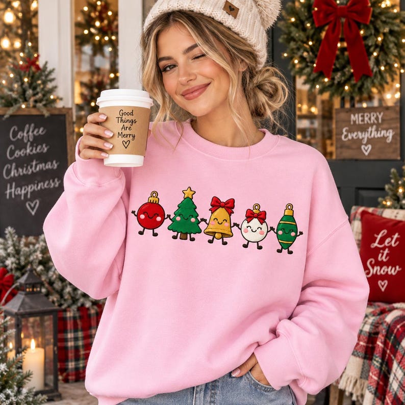 May include: A light pink crewneck sweatshirt featuring a graphic print of five cartoon Christmas ornaments holding hands, including a red bauble, a green tree, a yellow bell, a white bauble with a red bow, and a green teardrop ornament.