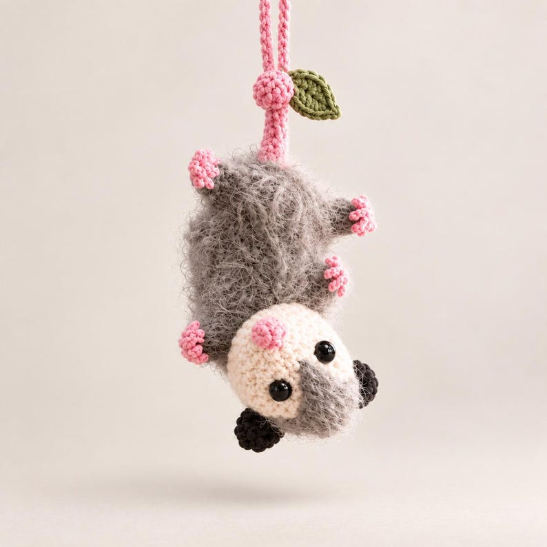 May include: A crocheted opossum hanging upside down by its pink tail. The opossum has a fuzzy gray body, a cream-colored face, black ears, and small pink feet. A small green leaf is attached to the base of the pink crocheted hanging loop.
