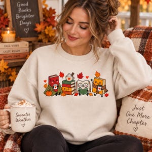 May include: A beige crewneck sweatshirt featuring a graphic of anthropomorphic books, a stack of books, and a mug with a heart. The design includes autumn leaves and a pumpkin. The sweatshirt is paired with a mug labeled Sweater Weather.